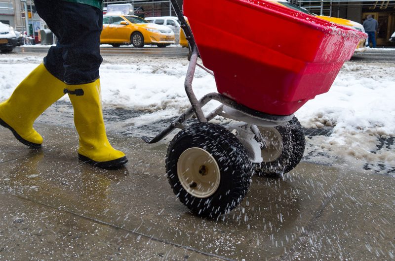 worker pushing salt spreader on icy New York City sidewalk during snow storm in winter.